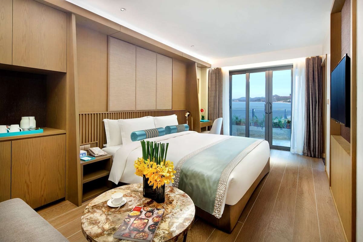 A hotel room with a large bed, a desk with a telephone, a coffee table with a magazine, and a view of the sea.