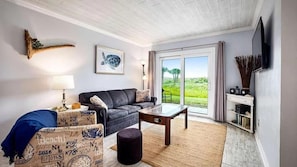 Uma sala de estar com sofá, mesa de centro, abajur, televisão e vista para o exterior.