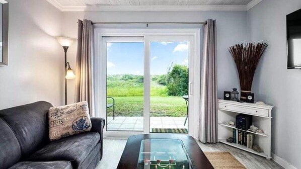 A living room with a sofa, a coffee table, a TV, and a bookshelf. There is a sliding glass door leading to a garden.