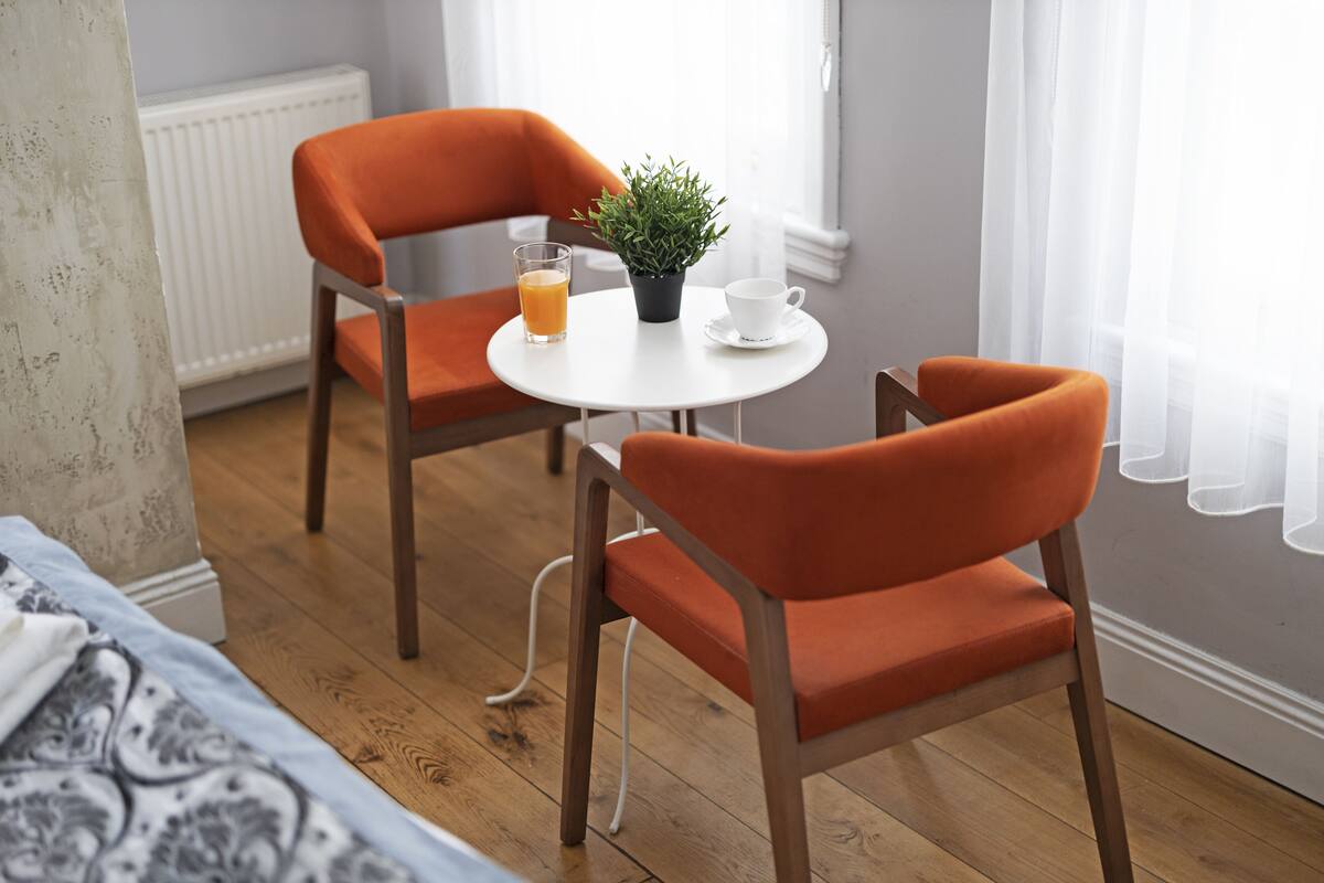 Una habitación con dos sillas naranjas, una mesa redonda pequeña, una planta en maceta, una taza blanca y un vaso de jugo de naranja.
