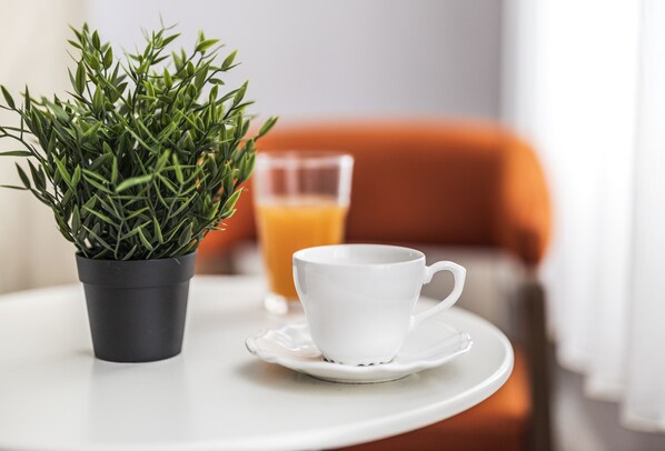 Une petite plante en pot, une tasse et une soucoupe blanches, et un verre de jus d’orange sur une table blanche.