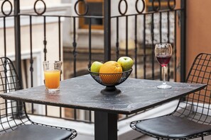 Une table avec un panier de fruits, un verre de jus d’orange et un verre de vin rouge, le tout sur un balcon.