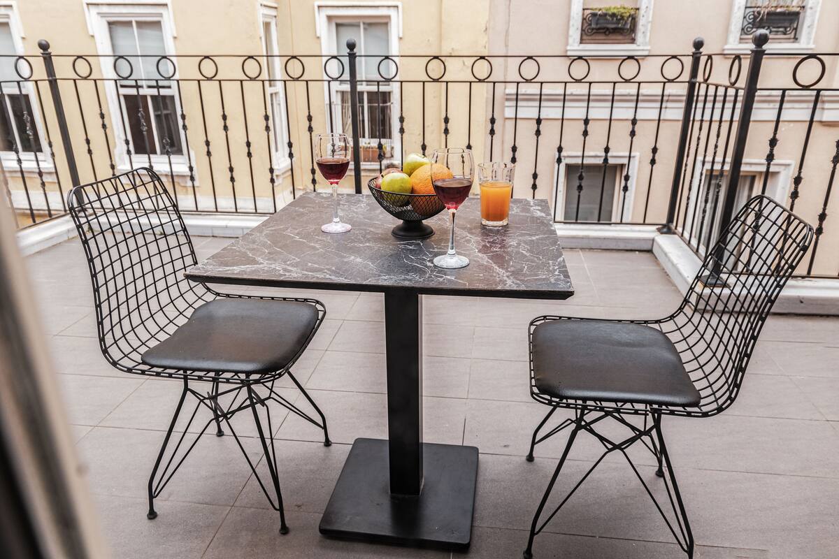 Un balcón con una mesa, sillas, un frutero y dos bebidas.