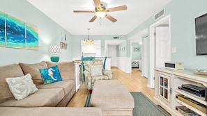A living room with a sofa, armchair, and a small table with books. There is a ceiling fan and a painting on the wall.