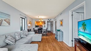 A modern living room with a grey sectional sofa, a dining area with a wooden table and chairs, a kitchen in the background, and a TV on the right.