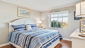 A bedroom with a large bed, a nightstand, a lamp, a framed picture, a window with a view, and a sign above the window.