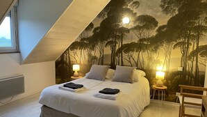 Ein Schlafzimmer mit schrägen Wänden, einem Bett mit weißen Bettwäsche, zwei Nachttischlampen und einem Wandgemälde von Bäumen.