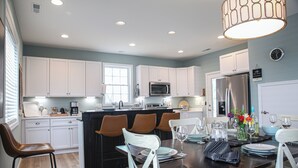 A modern kitchen with white cabinets, a central island, and a dining area with chairs and a table set for four. - NEW 3 BR/2.5BTH near Bethany Beach/home on stilts, open floor plan/1st FL Master (Ocean View)
