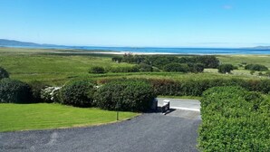Un paysage rural avec une allée de gravier menant à un bâtiment entouré d’une végétation luxuriante, et d’où l’on peut admirer la mer.
