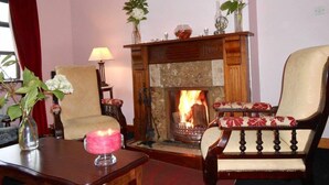 Un salon chaleureux avec une cheminée, deux fauteuils, une table ornée d’un vase et d’une bougie, et une lampe.