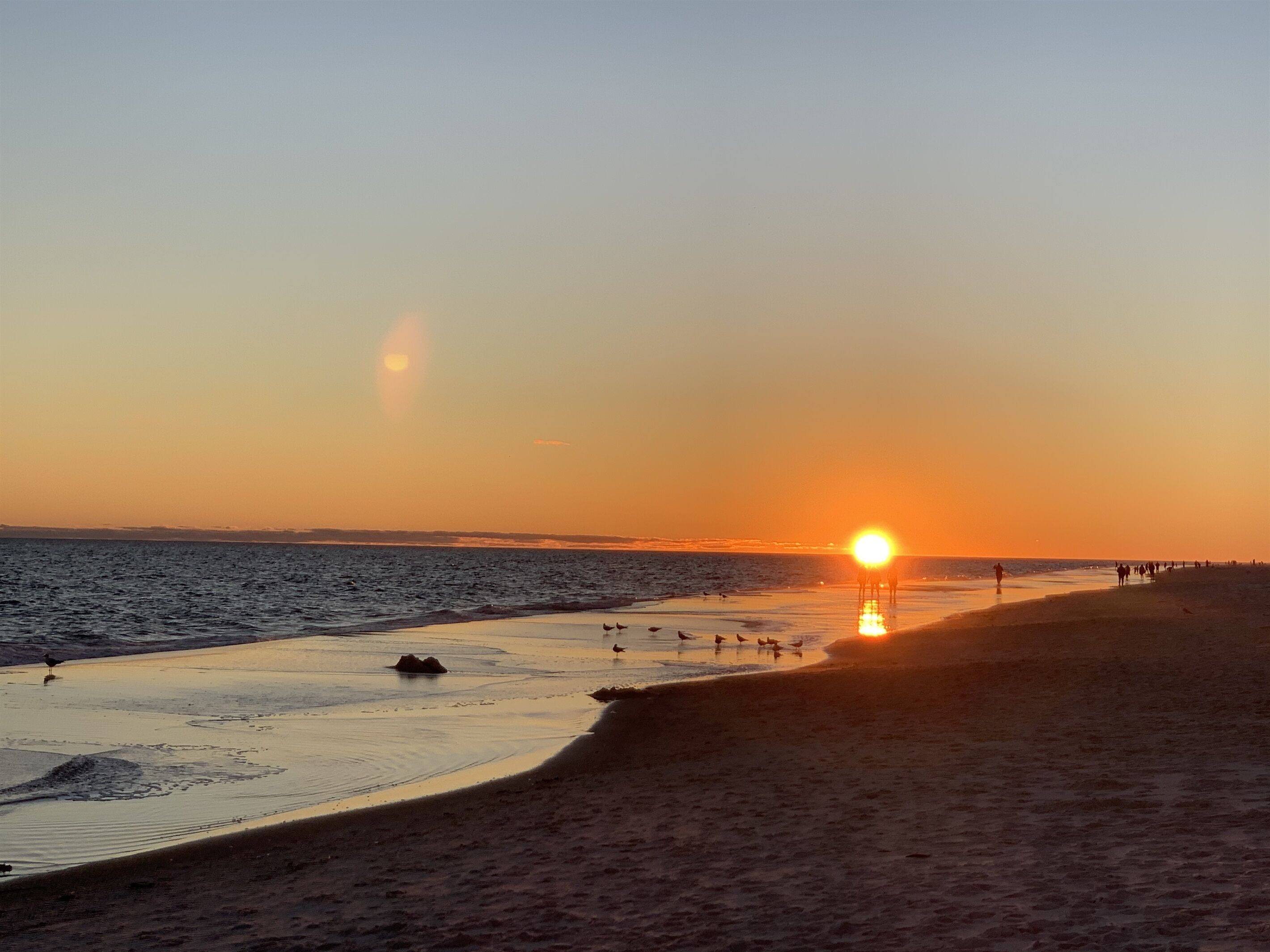 Une plage au coucher du soleil, avec le soleil bas à l’horizon, une mer calme et des personnes se promenant le long du rivage.