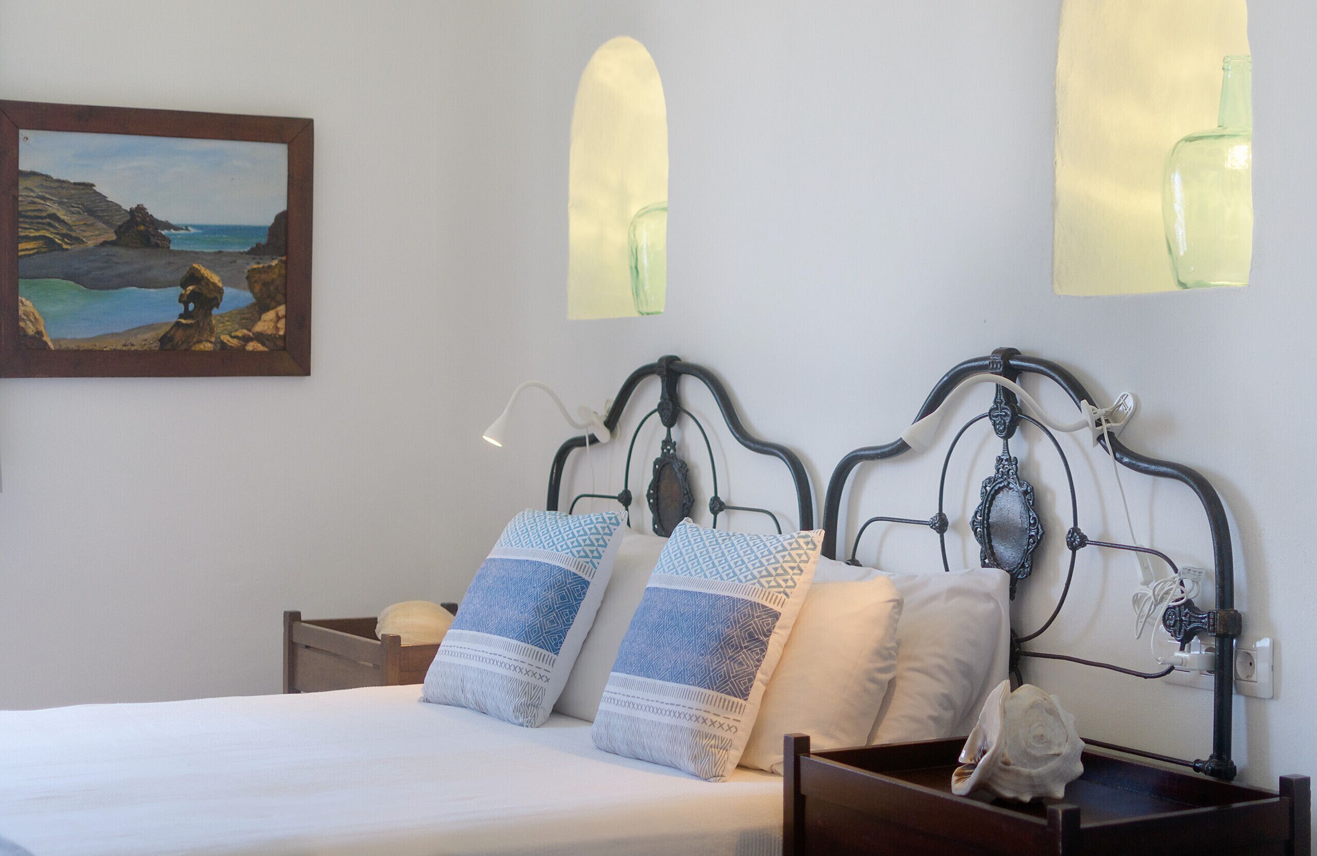 Cama bem arrumada com almofadas estampadas, cabeceira de madeira e pintura de uma paisagem costeira na parede.