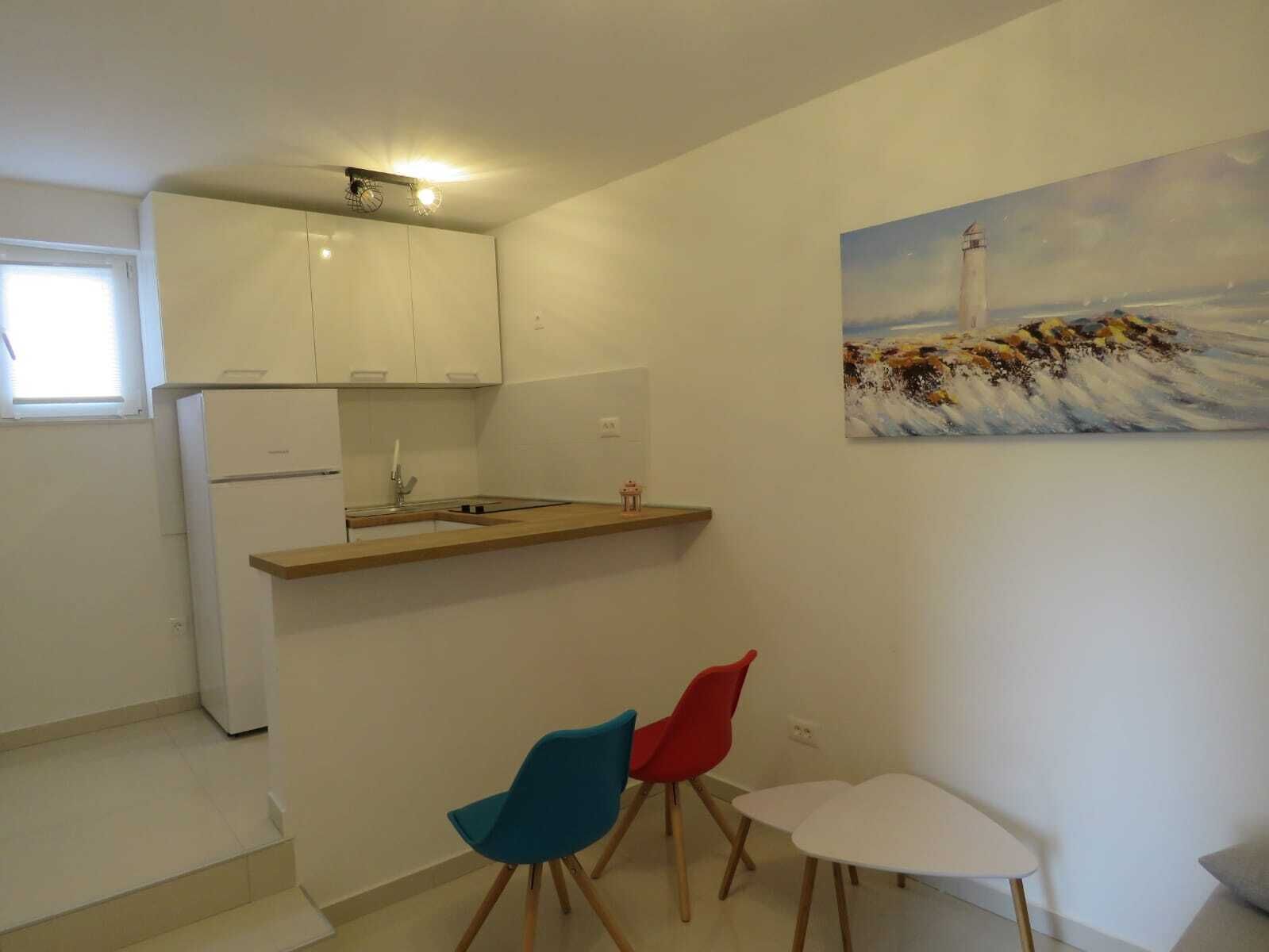 A compact kitchen with white cabinets, a small sink, and a wooden countertop. There are two chairs and a small white table. A painting of a lighthouse is on the wall.