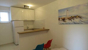Una cuina compacta amb armaris blancs, un petit rentapeus i un taulell de fusta. Hi ha dues cadires i una taula petita blanca. Al mur, hi ha un quadre d'un far.