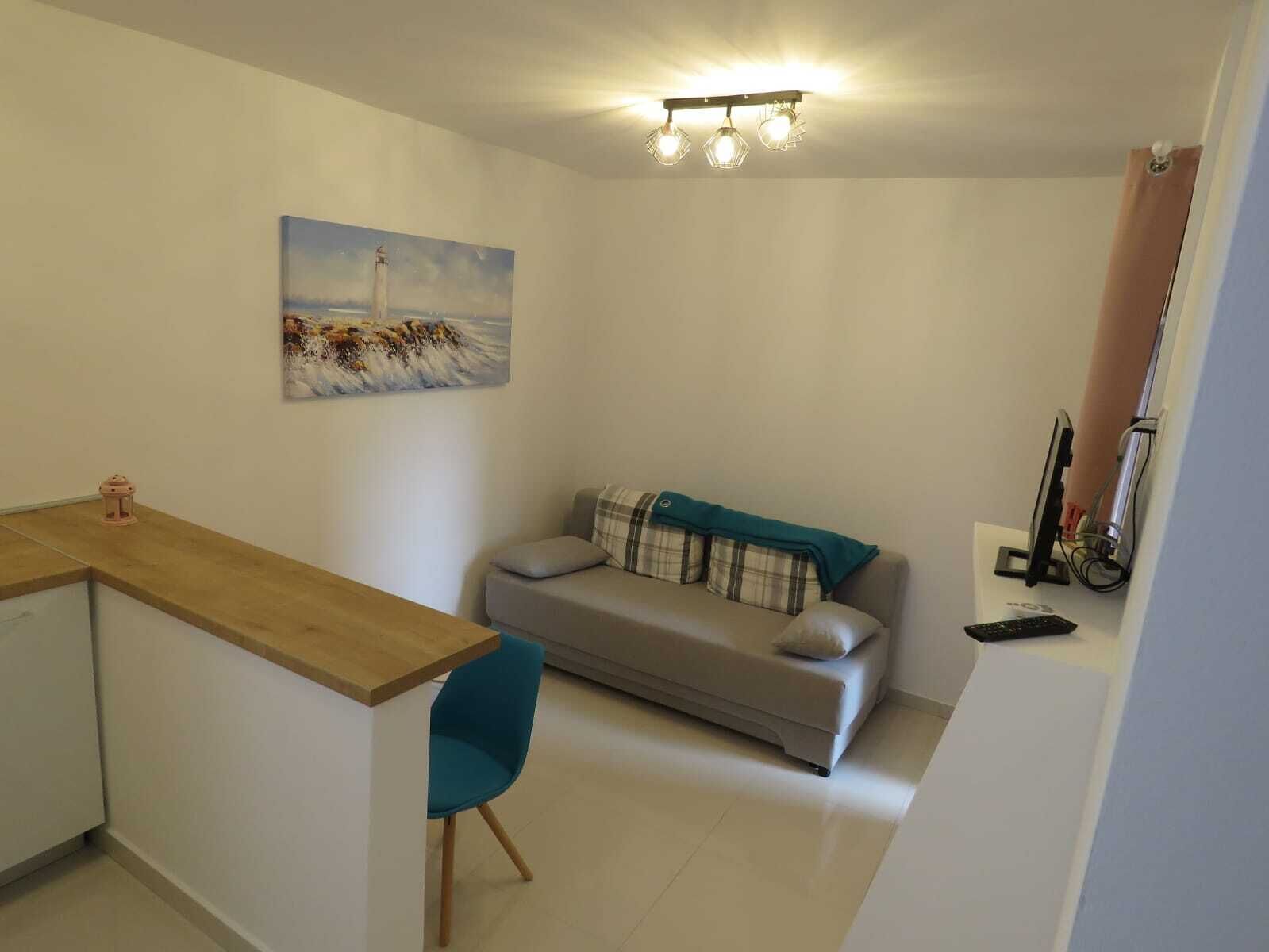 Un espace de vie compact comprenant un coin cuisine, un canapé avec un plaid, un fauteuil bleu et un tableau représentant un phare accroché au mur.
