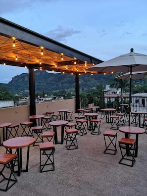 An outdoor seating area with tables and chairs, a pergola with string lights, and a view of a mountainous landscape.