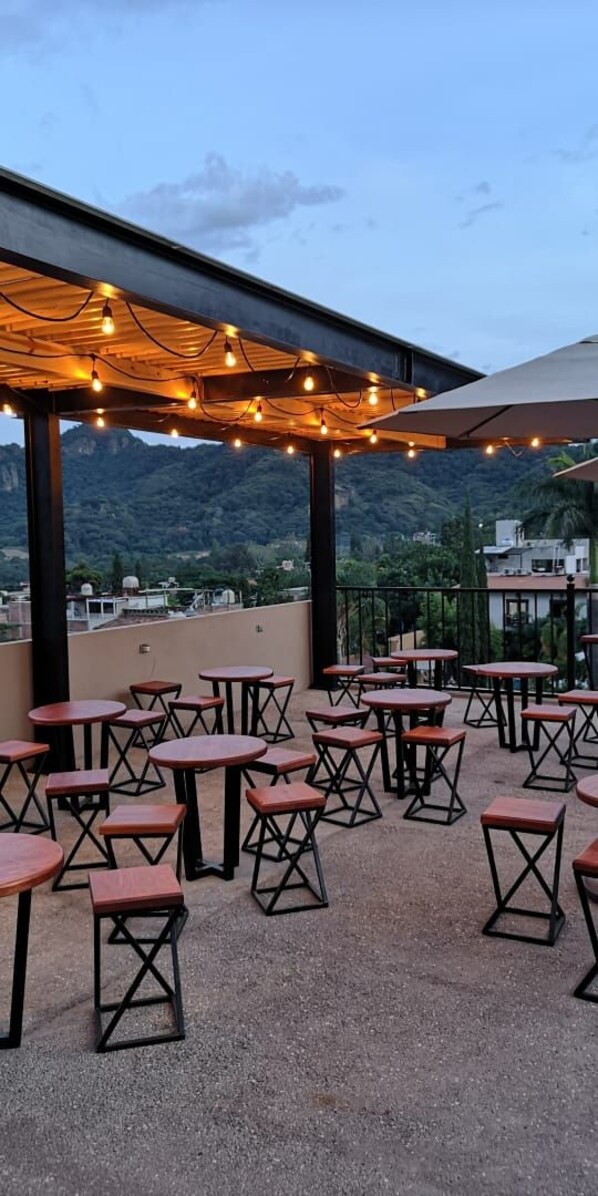 An outdoor patio with tables and chairs, a pergola with string lights, and a mountainous backdrop.