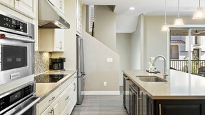 A modern kitchen with stainless steel appliances, a large island with a sink, and a view of the living area through an open door. - Luxury Rental by The Princess  (Scottsdale)