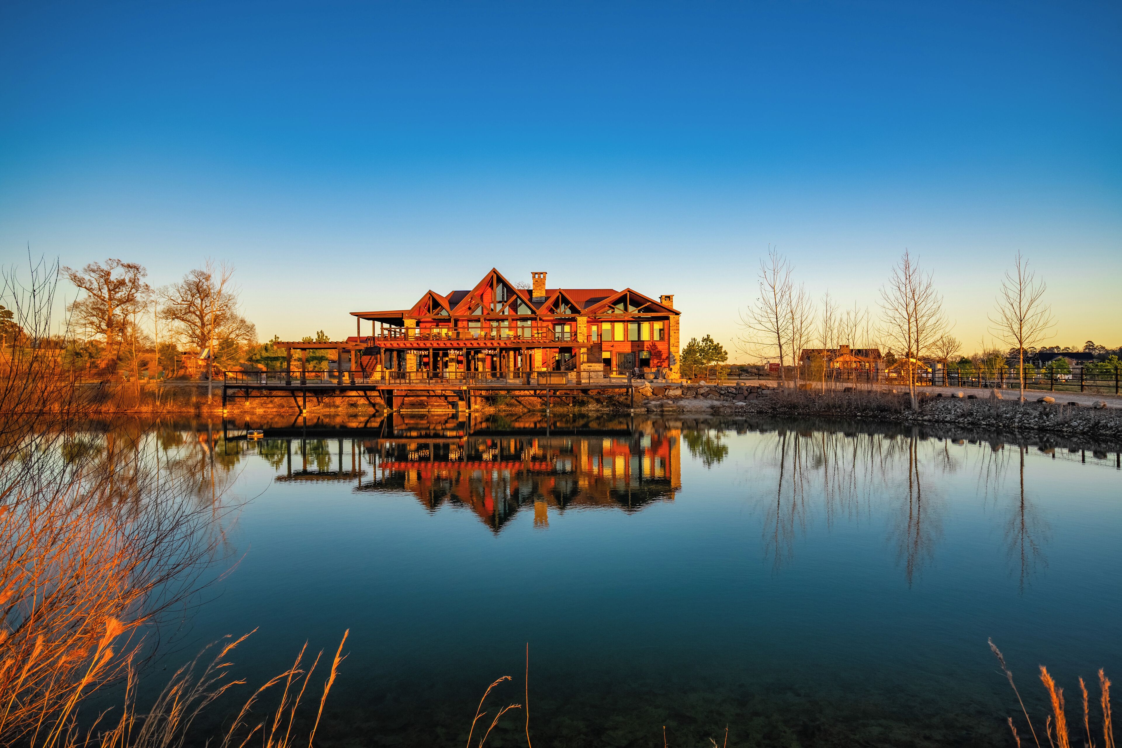 A large, multi-story house with a reflection in a calm body of water, surrounded by trees and a clear sky.
