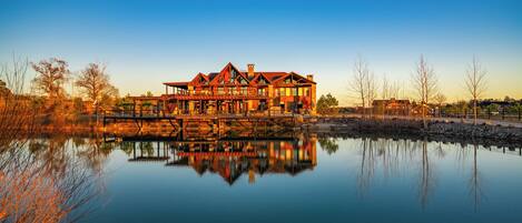 A large, multi-story house with a reflection in a calm body of water, surrounded by trees and a clear sky.