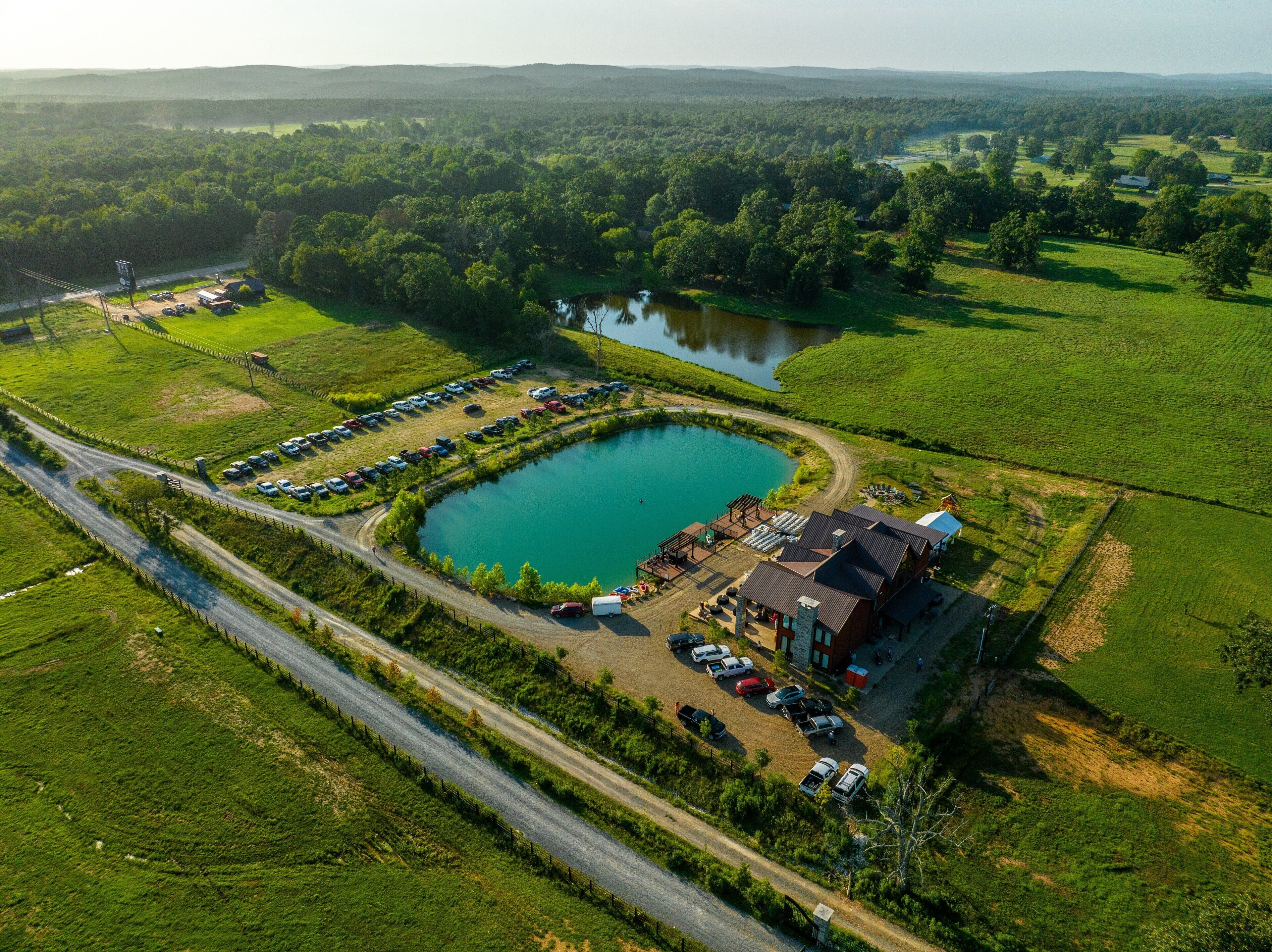 An aerial view of a rural property with a large pond, a parking area, and a building with a sloped roof.