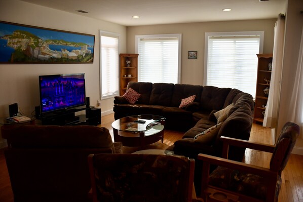 A living room with a large flat-screen TV, a dark brown sectional sofa, a glass coffee table, and a wooden bookshelf.