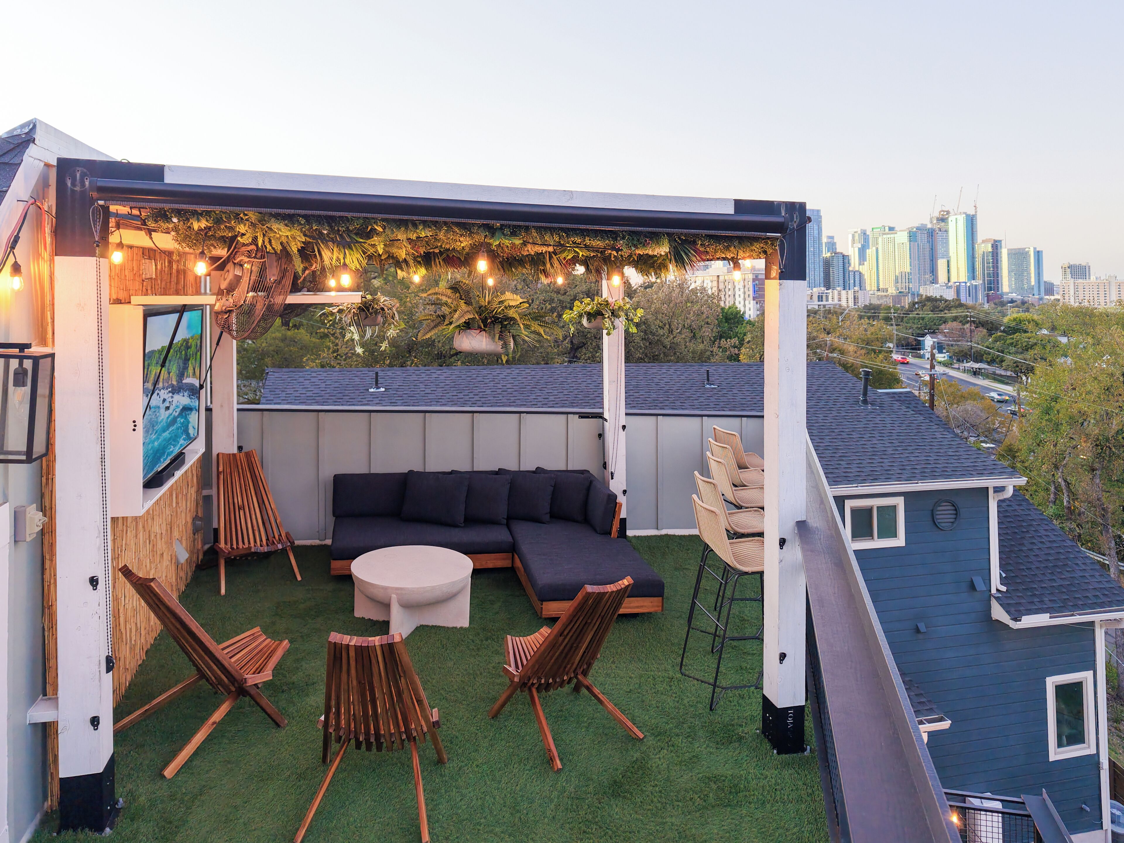 A rooftop patio with a sectional sofa, two wooden chairs, a round table, and string lights. A city skyline is visible in the background.
