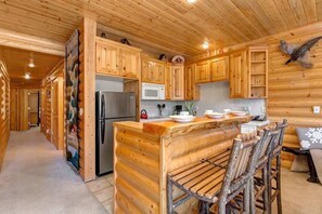 Innenansicht einer Blockhütte mit Küchenbereich, einschließlich Holzschränken, einem Kühlschrank, einer Mikrowelle und einem Barhocker.