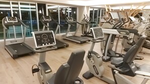 A gym with multiple treadmills and exercise bikes, large windows, and a wooden floor.