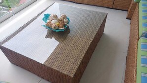 A room with a large, rectangular, woven-texture coffee table with a glass top, a blue ceramic bowl with decorative items on it, and wicker furniture in the background.
