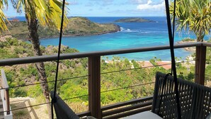 Ein Balkon mit einer Hängematte, der auf eine Küstenlandschaft mit klarem, blauem Wasser und grünen Hügeln blickt.