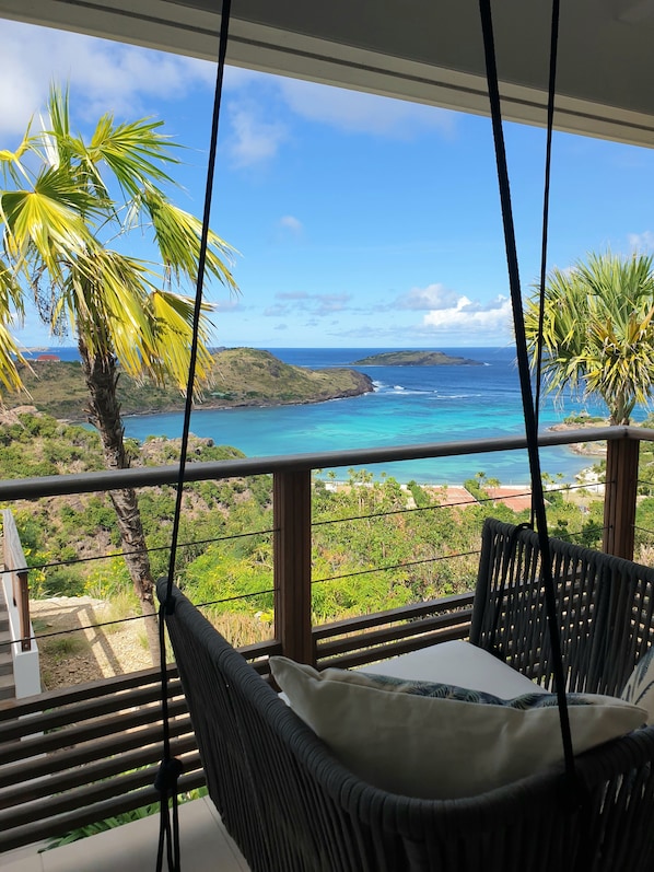 Un balcone con una poltrona sospesa che si affaccia su un paesaggio costiero con acque blu cristalline e colline verdi.