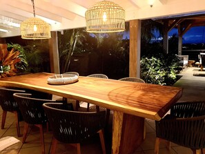 Un espace de restauration en plein air avec une table en bois et des chaises en osier, entouré d’une végétation luxuriante et offrant une vue sur le ciel nocturne.