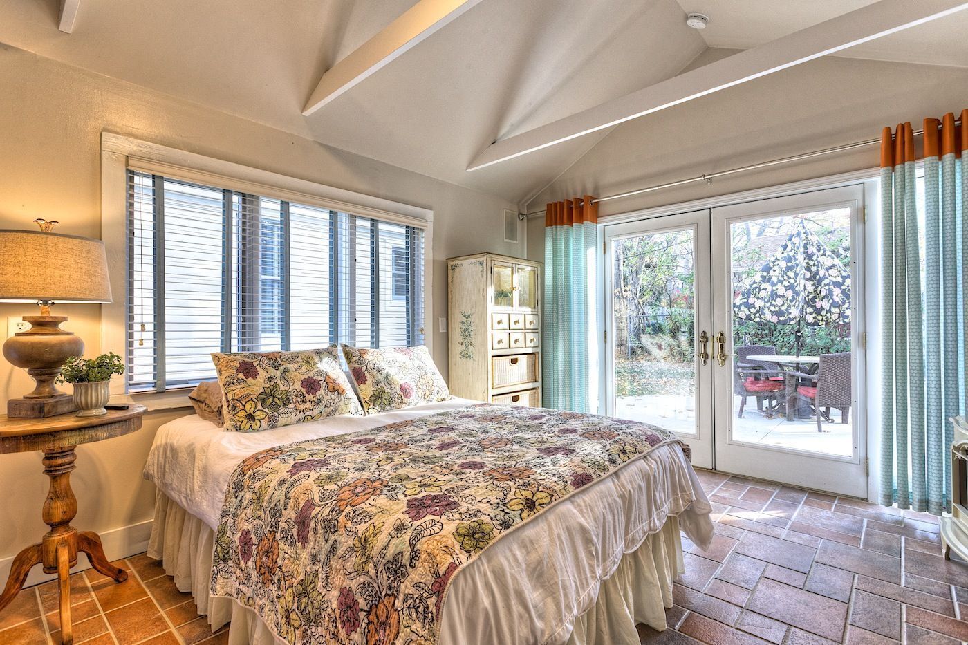 Une chambre avec un grand lit, une table de chevet, une lampe, une fenêtre avec des stores, une armoire et une porte-fenêtre coulissante donnant sur un espace extérieur.