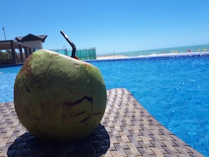 A large green fruit with a straw on a woven table by a pool with a clear view of the beach. - Riviera Beach Place Golf Residence (Aquiraz)