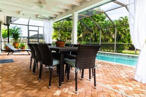 Un patio al aire libre con mesa y sillas, una piscina y abundante vegetación de fondo.