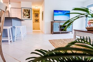 Ein modernes Wohnzimmer mit Fernseher auf einem Holzkabinett, ein Küchenbereich mit weißen Barhocker und ein Flur mit gerahmten Bildern.