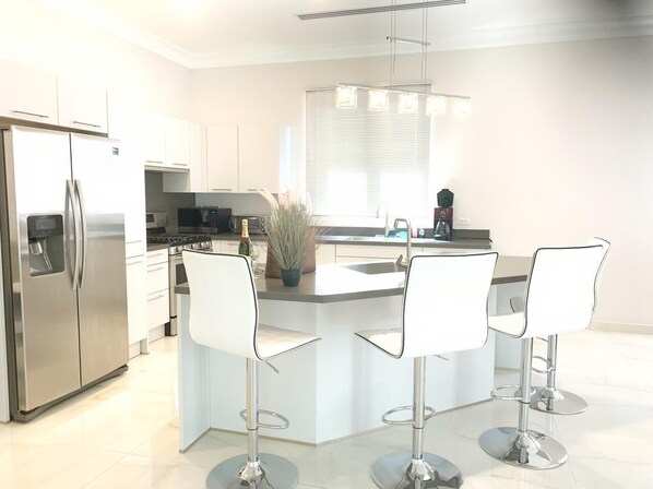 Een moderne keuken met een centraal eiland, roestvrijstalen apparatuur en witte barkrukken.