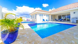 Une piscine avec une terrasse carrelée, un espace patio aménagé avec du mobilier d’extérieur et une maison avec un toit recouvert de tuiles.