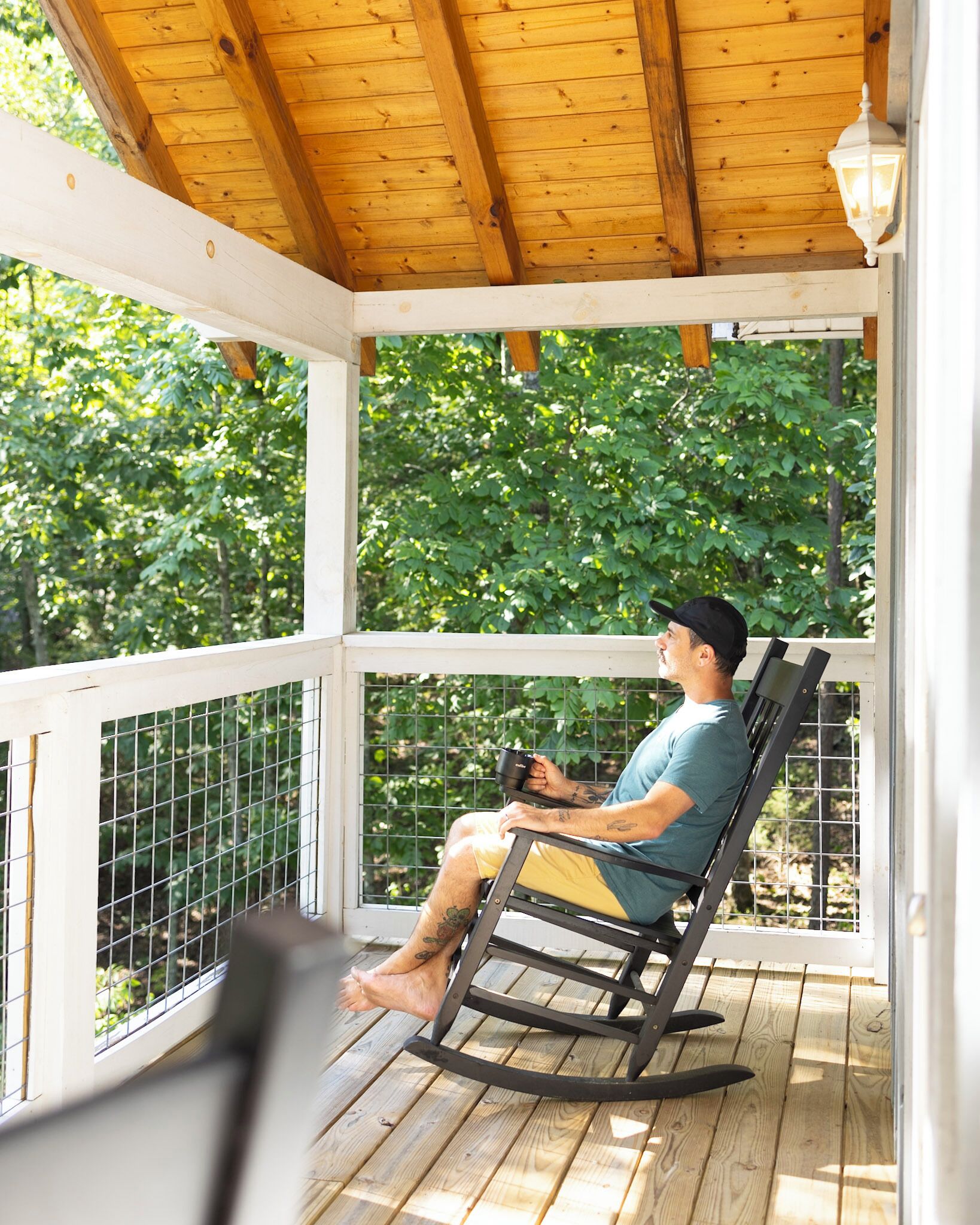 A man sitting on a rocking chair on a wooden deck with a metal railing, overlooking a garden with green foliage.
