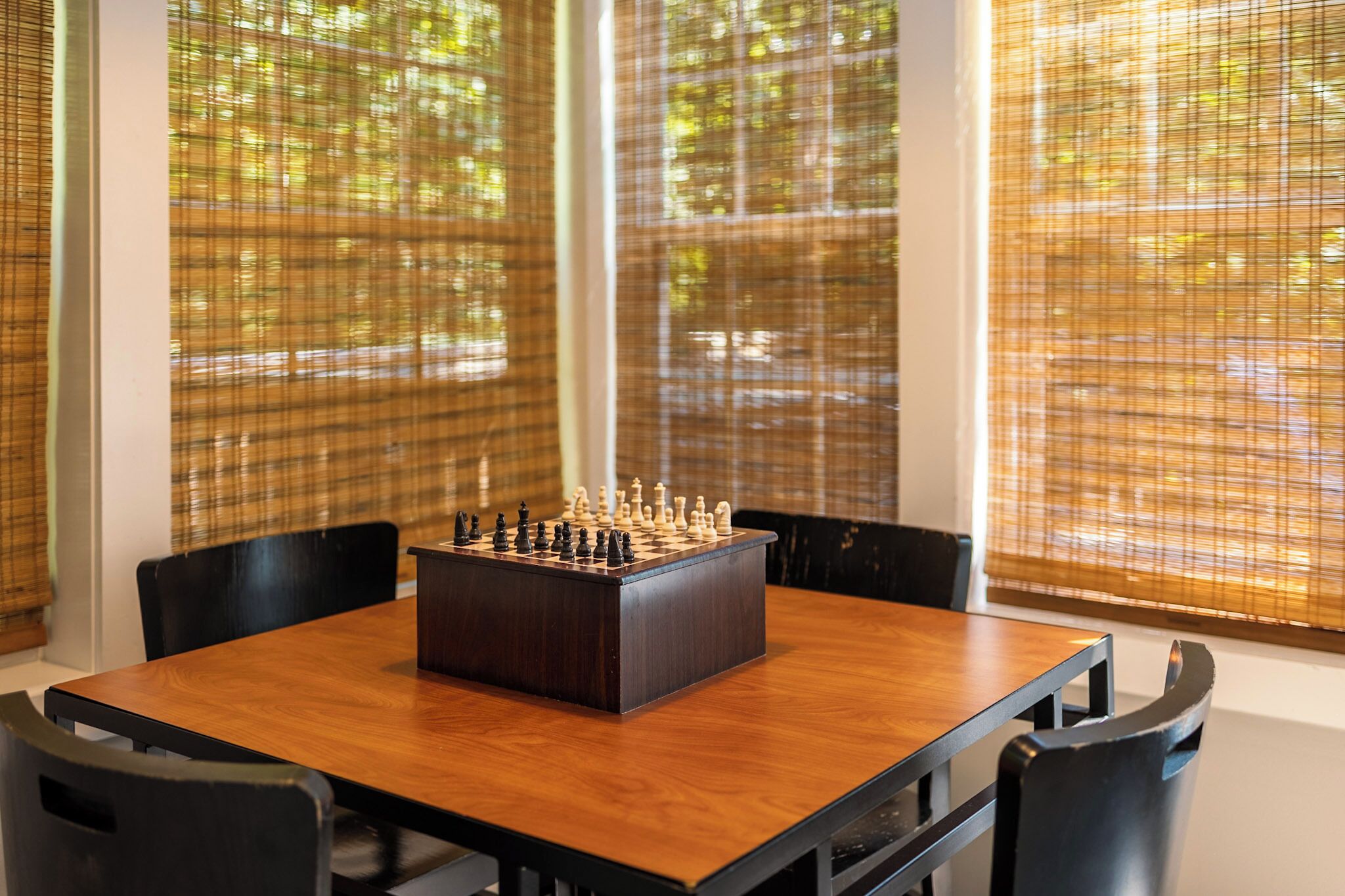Un échiquier posé sur une table en bois, dans une pièce où se trouvent des chaises noires et de grandes fenêtres équipées de stores.