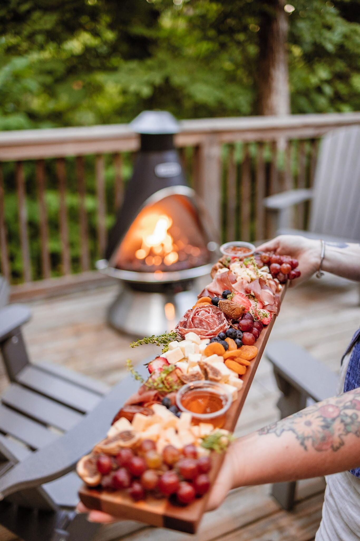 A person holding a long wooden board with various cheeses, fruits, and meats, with a fire pit in the background.