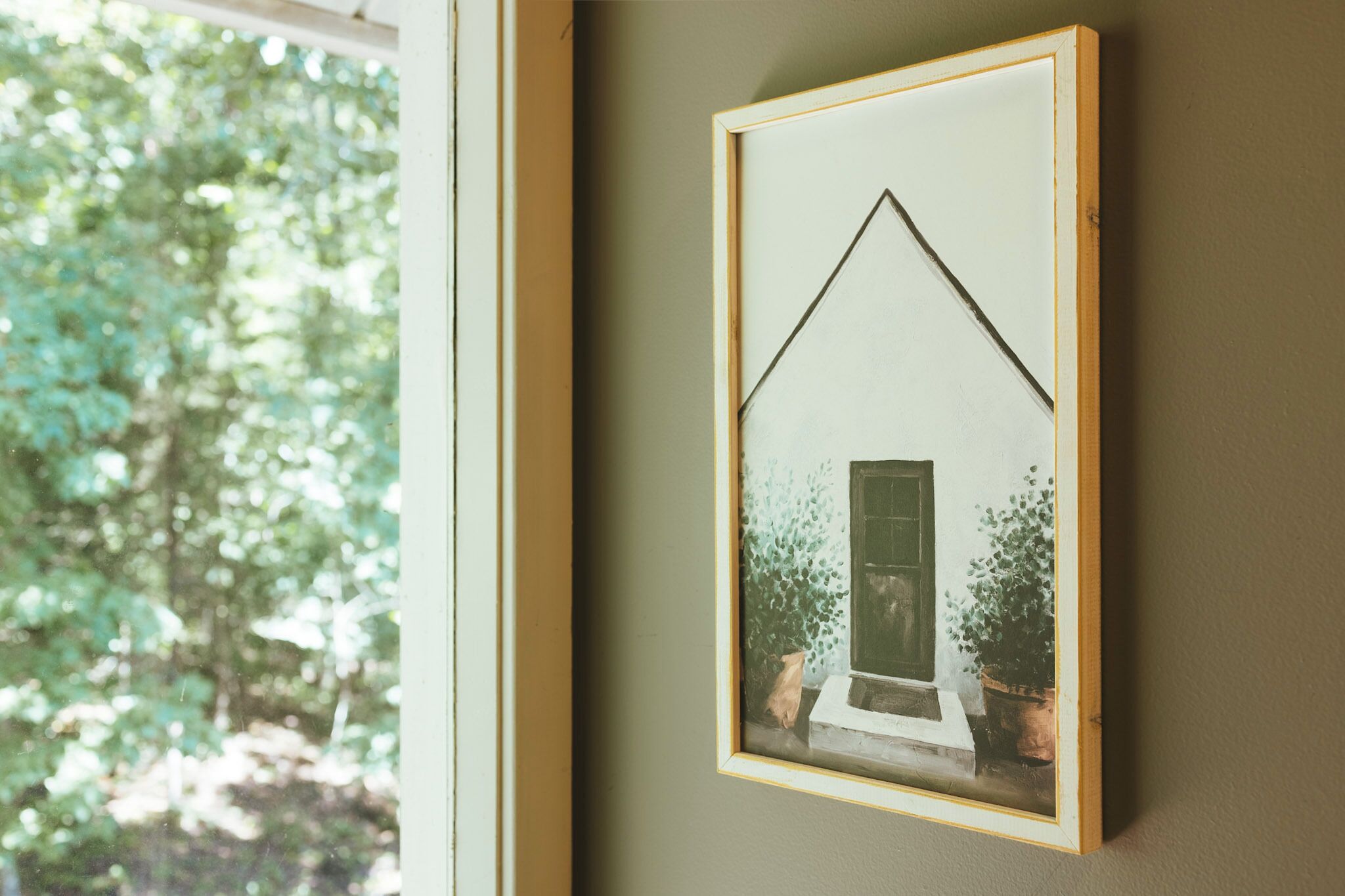 A framed picture of a house with a triangular roof, potted plants, and a door on a wall.