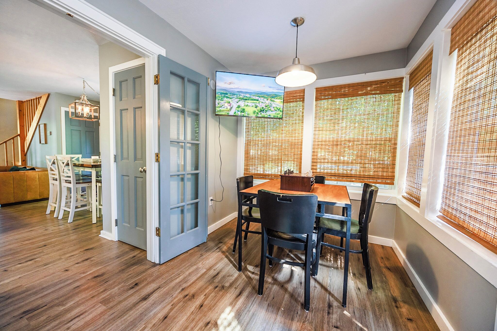 A dining area with a wooden table and chairs, a television mounted on the wall, and a window with bamboo blinds.