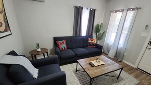 A living room with a dark sofa, a coffee table, a small side table with a plant, and a window with curtains. - WL1- Modern 2BR Retreat Minutes from Downtown & Top SA Attractions (San Antonio)
