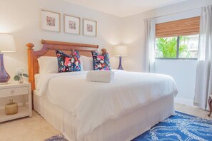 A neatly made bed with white linens, a wooden headboard, and floral pillows. There are two bedside tables with lamps and a small vase of flowers. A window with curtains and a view of greenery is visible.