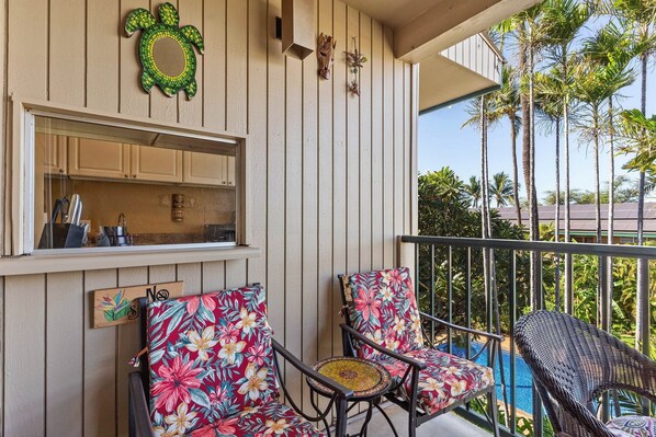A balcony with a pool, two chairs with floral upholstery, a woven chair, and a kitchen area with cabinets and a window.