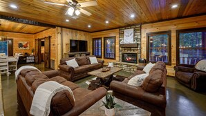 A cozy log cabin interior with wooden walls, a fireplace, leather sofas, and large windows. - THE PORCH AT BROKEN BOW 5b/6ba/Sleeps 20 - 2pm Checkout! Loads of amenities! (Broken Bow)