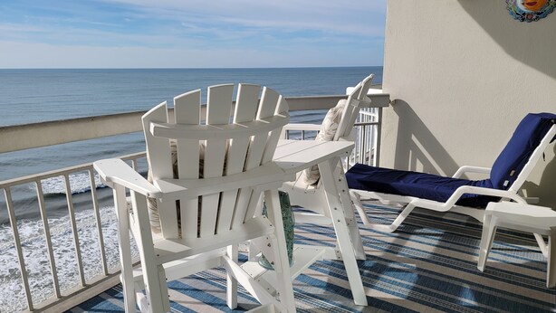 Un balcon agrémenté de chaises Adirondack blanches et d’un coussin bleu, donnant sur une plage où l’on peut admirer les vagues.