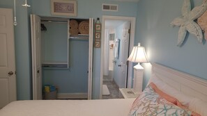 A bedroom with a bed, a nightstand, a lamp, a closet, and a hallway leading to a bathroom.